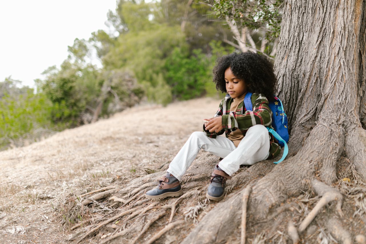 A young boy with curly hair sits against a tree outdoors, wearing a plaid shirt and backpack, enjoying nature.